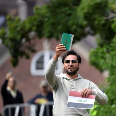 En man med solglasögon håller upp en bok med grön pärm i ena handen och en bild av Iraks flagga i andra handen.