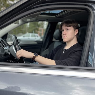 Nuori autonkuljettaja istuu auton ratissa auton ikkuna auki, poika on pukeutunut mustaan.