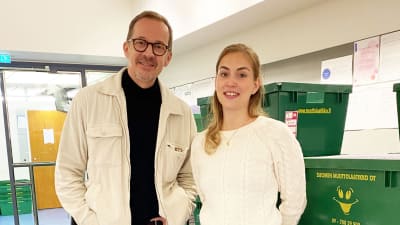 Två personer står i en korridor där det finns staplar med flyttlådor längs väggarna. De ser in i kameran och ler.