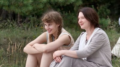 Malin (Saga Sarkola) och Salome (Satu Tuuli Karhu) sitter bredvid varandra på en strand och ler.