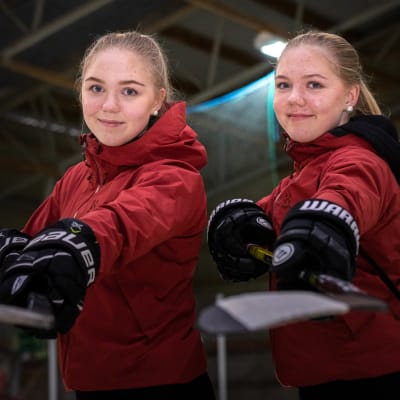 Iida (vas.) ja Emma Lappalainen punaisissa takeissa, jääkiekkohanskat kädessä, ojentamassa jääkiekkomailoja kameraa kohti.