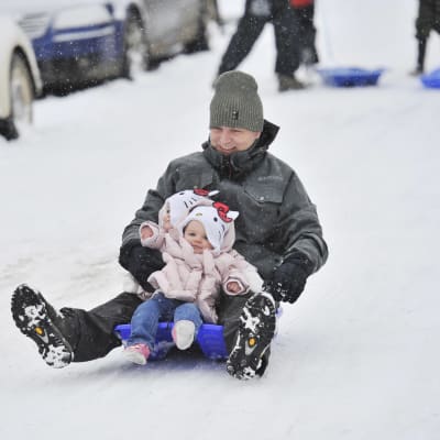 En man håller i ett litet barn medan de åker pulka ner för en backe.