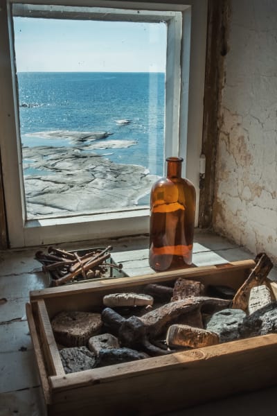 Fönsterbräde med brun glasflaska, låda med stenföremål och ask med rostiga spikar. Utsikt över hav i solsken.