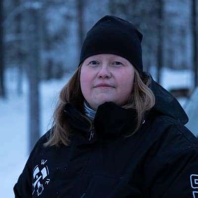 Tiina Sanila-Aikio katsoo totisena kameraan. Päällään musta takki ja musta pipo. Taustalla näkyy luminen maisema jossa poroja ja moottorikelkka..