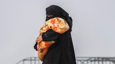 En kvinna i niqab med ett barn i famnen promenerar med vita tält i bakgrunden.