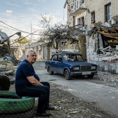 En ädra man sitter på ett bildäck, i bakgrunden en blå Lada och ett utbombat hus.