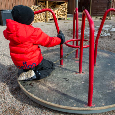 Ett barn i tjock röd jacka på en karusell i en lekpark. 
