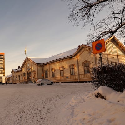 Talvinen kuapunkimaisema Oulun asema-aukiolta. Keltainen vanha VR:n puinen rautatieasema ja sen takana ruskea kerrostalo. Tien toisella puolella asemaa vastapäätä kerrotaloja ja autoja.