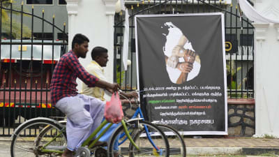 Cyklister på Sri Lanka vid en plansch som visar fyra händer med olika religiösa symboler