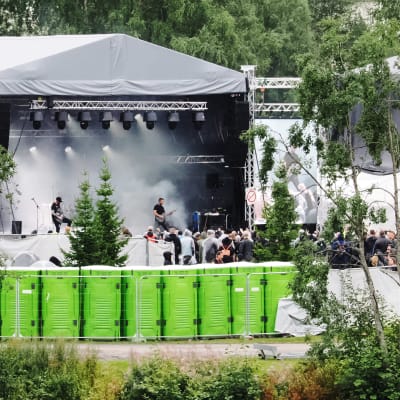 Rock bändi soittaa esiintymislavalla savutehosteiden keskellä ja edessä sadevarusteisiin pukeutunutta yleisöä. Etualalla rivissä vihreiden käymälöiden takaseiniä.