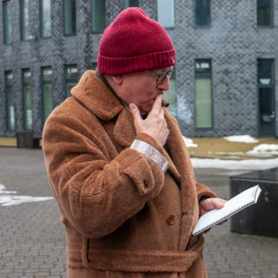 En äldre man i beige yllekappa och röd ylleluva röker cigarr och tittar på ett papper.