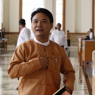 Mies, Phyo Zeya Thaw, seisoo Myanmarin parlamentin salissa, päällään perinteinen takki, käsi napituksen aukossa. 
