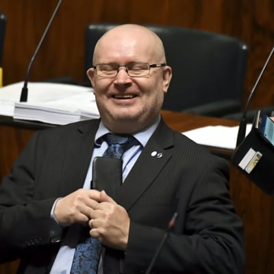 En skallig man i kostym med en telefon i handen skrattar i riksdagen.