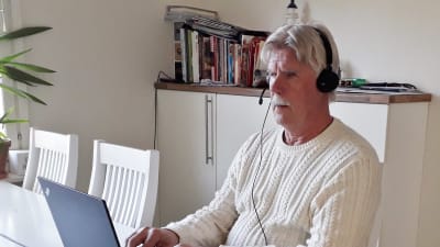 En man i övre medelåldern har headset på huvudet och sitter vid sitt köksbord framför sin laptop och skriver.