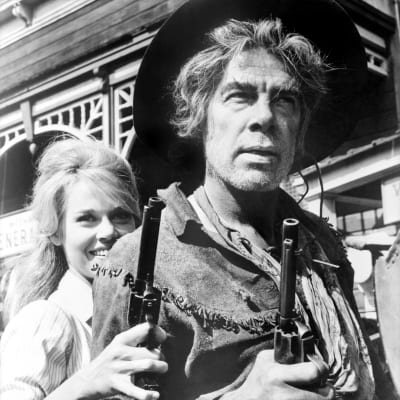 Hattupäinen, resuisen näköinen lännenmies (näyttelijä Lee Marvin) takanaan hiukset auki hymyilevä nainen (näyttelijä Jane Fonda), molemmilla kädessään iso reikärauta, kuva elokuvasta Paukkurauta-Kati.