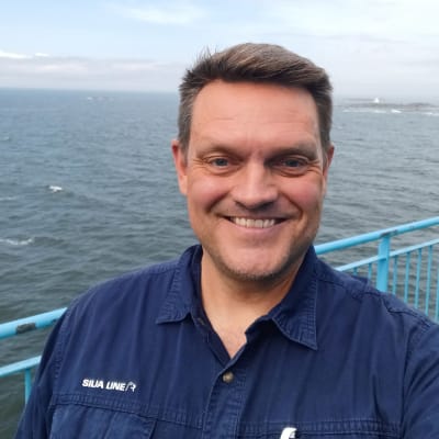 En man med mörkt hår, ett stort leende och en blå overall med texten Silja Line står på ett fartygsdäck i skärgården.