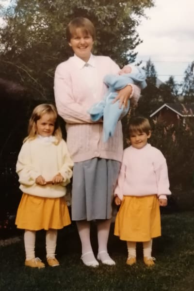 Ett gulnat fotografi av en kvinna. I famnen håller hon ett spädbarn och bredvid henne står två flickar i gula kjolar. 