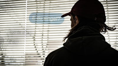 En man i 20-årsåldern står med ryggen mot kameran och tittar ut genom ett fönster med spjälgardiner. Stämningen är hotfull. Det är en dramatiserad bild som föreställer en nätbedragare.