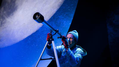 En man i mössa och vantar tittar i en stjärnkikare. I bakgrunden syns en väldig måne. 