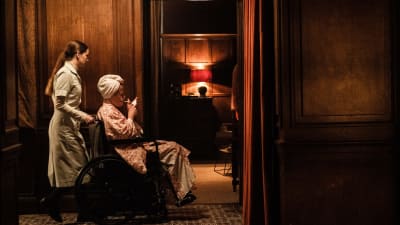 Maud skuffar fram en rökande Amanda som sitter i rullstol.