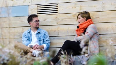 Adam Guarnieri och Maria Sundblom Lindberg i samspråk utomhus framför ljus trävägg på ön Lonnan i Helsingfors.