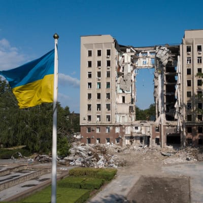 En administrationsbyggnad i Ukraina är sönderbombad i mitten. I förgrunden syns en ukrainsk flagga.