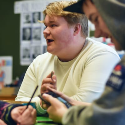 En und man med ljust hår och vit tröja pratar och ser glad ut. Bredvid honom syns en ung man med luva och keps i profil.
