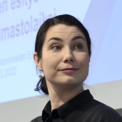 En mörkhårig kvinna framför en stor bildskärm.