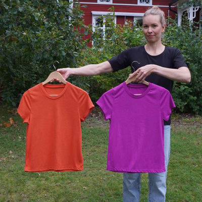 En kvinna som står i en trädgård framför ett rött hus. Kvinnan håller i två t-skjortor.