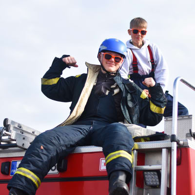 Två pojkar i brandkårskläder och solglasögon sitter på en brandbil.