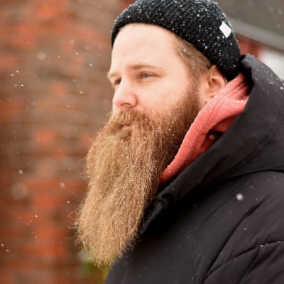 Man med långt ljus skägg iklädd svart mössa och jacka. Han står framför en tegelbyggnad. Det snöar.