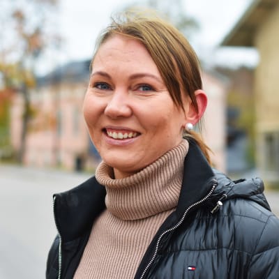 En kvinna i beige polotröja och svart jacka ser in i kameran och ler. Hon är på en gata utomhus.