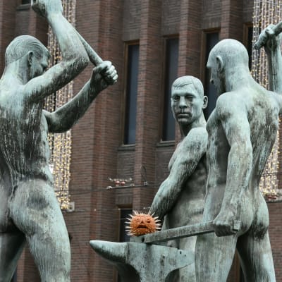 Skulpturen Tre smeder utanför Stockmann i Helsingfors. Under smedernas hammare har någon lagt en modell av ett coronavirus.