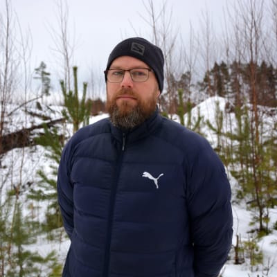 Man i 40-årsåldern med skägg och glasögon och mössa på huvudet står utomhus och tittar in i kameran.