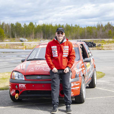 Rallisprint kuljettaja Oskari Heikkinen Ford-merkkisen punaisen ralliautonsa edessä.
