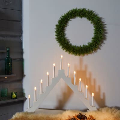 Ett julpyntat hörn i ett hem där en tunn tallriskrans hänger på en vit vägg, en stor adventsljusstake står tänd och några små lyktor lyser på sidan.