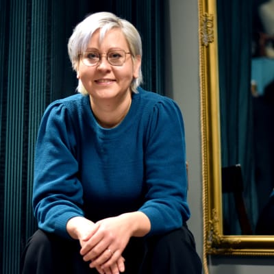 Kvinna med ljust hår och glasögon iklädd en blå tröja och svarta byxor sitter på en stol. En spegel och ett blått skynke i bakgrunden. Hon ler.