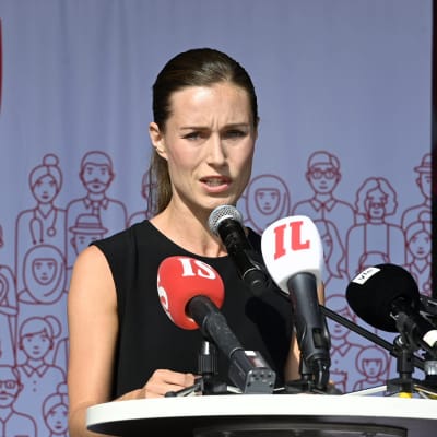 Statsminister Sanna Marin håller tal framför en rad med mikrofoner med SDP:s logotyp på en duk bakom sig.