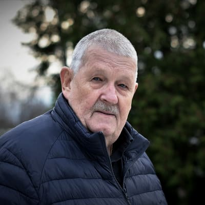 En äldre man står utomhus i novembermiljö och ser rakt in i kameran.