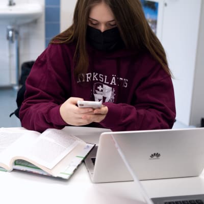 En elev scrollar på sin telefon. Framför sig har hon en dator och en uppslagen bok.