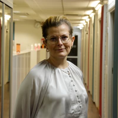 Kvinna med glasögon och kort hår i 40-årsåldern står i en korridor och tittar in i kameran.