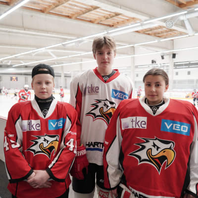 tre unga hockeyspelare med hockeyutrustning på står bredvid rinken och tittar in i kameran.