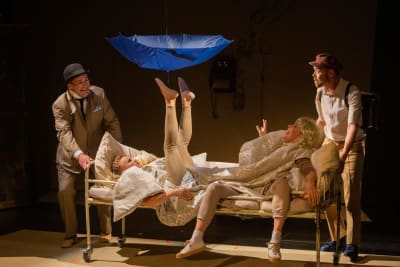 En grupp skådespelare står kring en säng, och i sängen lyfter en av artisterna upp ett paraply med fötterna.
