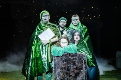 På en mörk teaterscen står fem skådespelare nära varandra. Alla är klädda i gröna klädesplagg. I bakgrunden syns stjärnor.