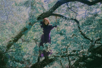 Säveltäjä, muusikko Astrid Swan seisoo puun oksalla lehvien siimeksessä violetissa mekossaan.