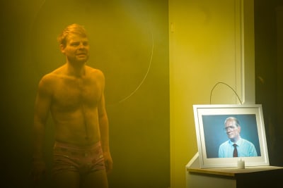 En barbröstad kille står i gult ljus bredvid ett inramat fotografi av Ebbe Carlsson. 