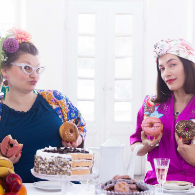 Kuvataiteilija Jasmin Anoschkin kirjavissa vaatteissa ja kukkia hiuksissaan sekä valokuvaaja Meeri Koutaniemi purppuranpunaisessa puserossa huivi päässä istuvat kakkuja, leivoksia ja hedelmiä notkuvan pöydän takana ja katsovat kameraan keikistellen ja pidellen kuorrutettuja donitseja rintojensa edessä.
