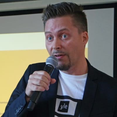Ville Grönberg puhuu mikrofoniin, taustakankaalla Mun vuoro -tiimin esitys