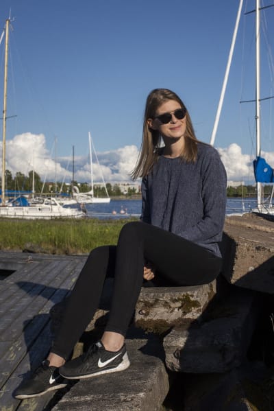 Sara Jakobsén sitter på en brygga. Hon har blå tröja och svarta byxor. I bakgrunden syns segelbåtar. 