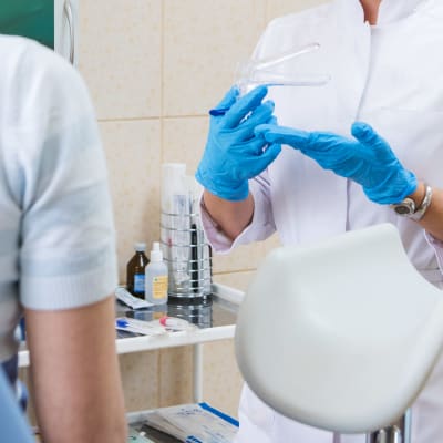 En läkare drar på sig blå plasthandskar medan en kvinna som man endast ser ryggen av sitter i en gynekologisk undersökningsstol.